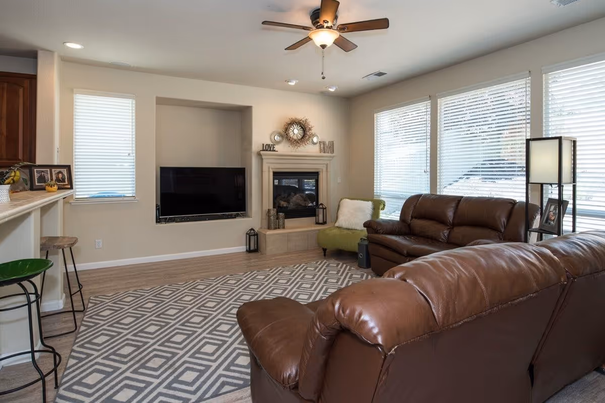 A cozy living room with two brown leather sofas, a green armchair with a white pillow, a patterned area rug, a wall-mounted flat-screen TV, and a fireplace decorated with a clock and small ornaments. Large windows with blinds allow natural light to fill the room, and a ceiling fan with a light fixture is mounted on the ceiling.