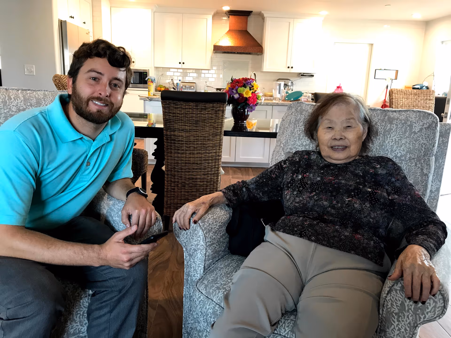 An elderly woman smiling in an armchair next to a younger man in a turquoise polo, with an open kitchen and dining area behind them.