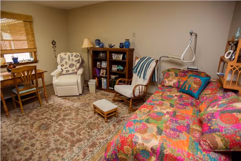 Cozy senior living bedroom with a colorful daybed, armchairs, a small bookcase, desk, and patterned rug.