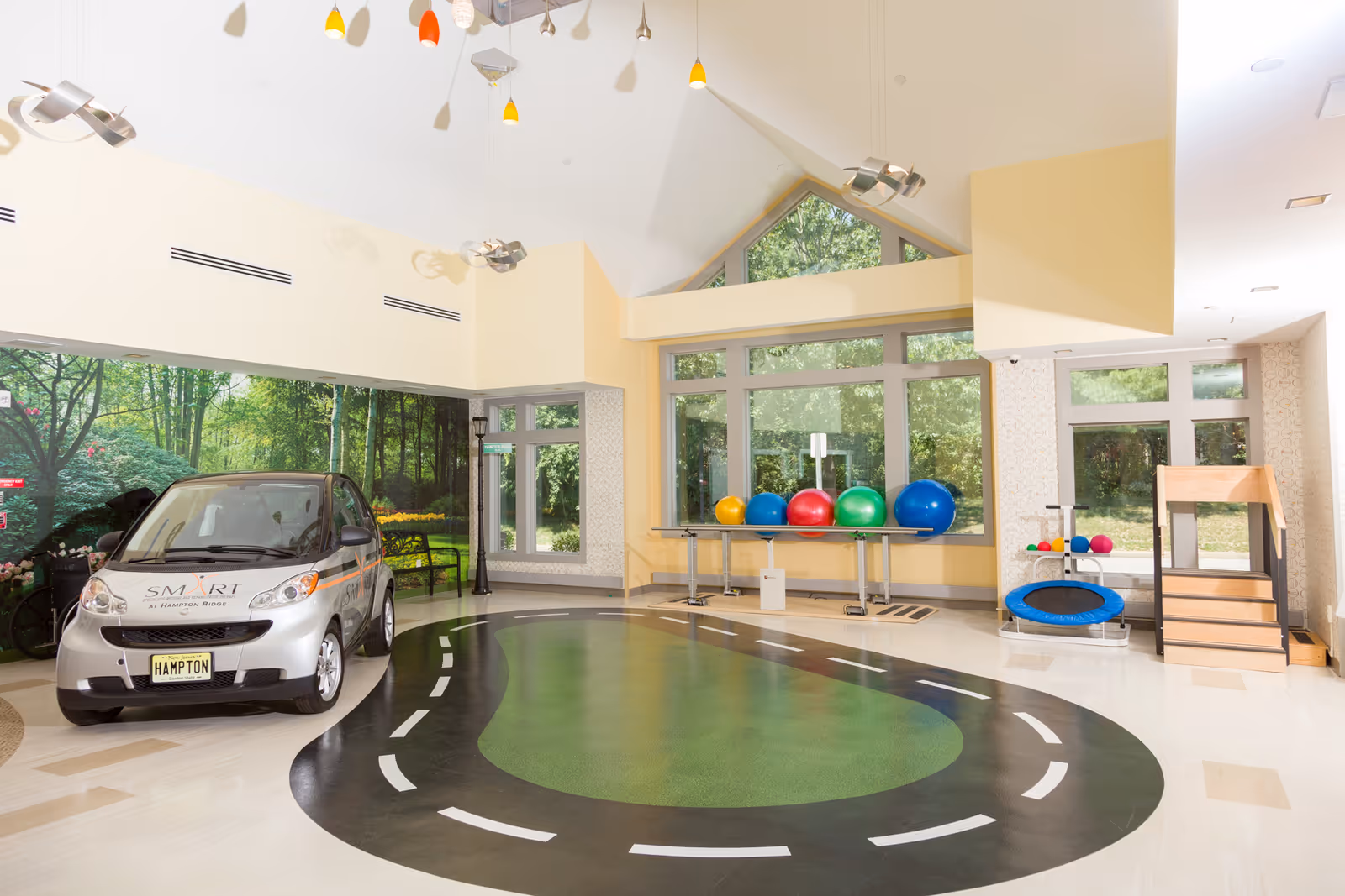 Indoor therapy or activity room with a small Smart car labeled 'SMART at Hampton Ridge' parked on a floor painted with a road track design. The room has large windows showing greenery outside, colorful exercise balls on a table, a small trampoline, and wooden stairs for physical therapy or exercise.
