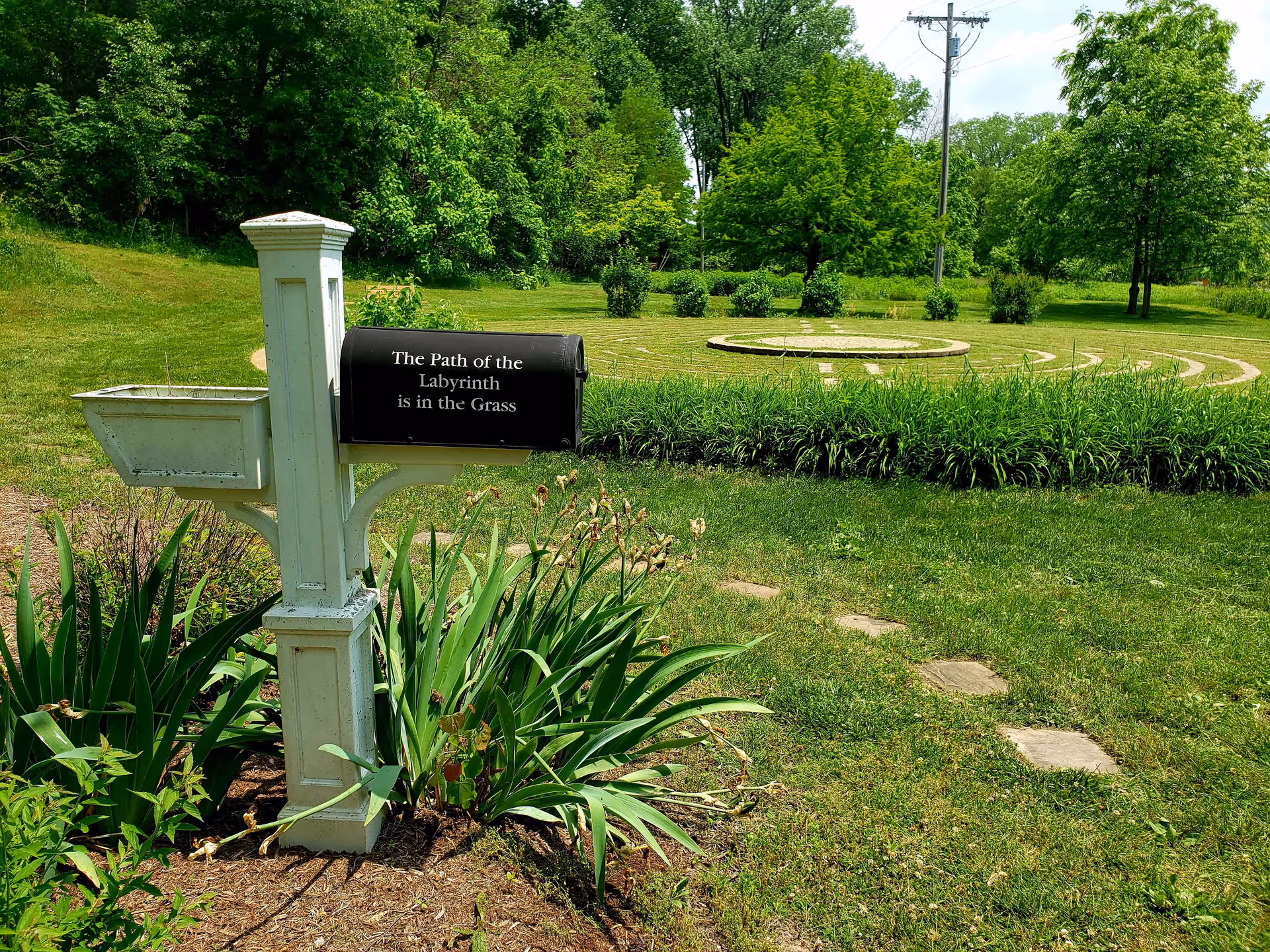 A white mailbox post with a black mailbox that has the text 'The Path of the Labyrinth is in the Grass' on it, surrounded by green plants and grass. In the background, there is a circular labyrinth pattern made of stone set in the grass, with trees and bushes further behind under a partly cloudy sky.