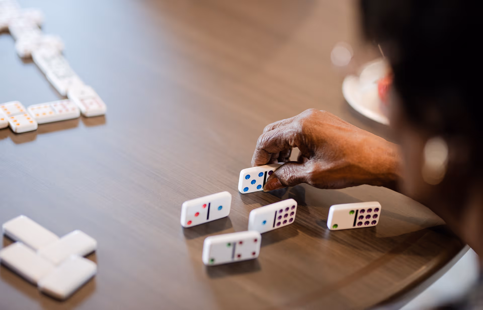 Close-up of a person's hand placing a domino piece on a wooden table with several other dominoes arranged on the table.