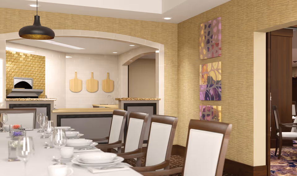 A dining room with a long table set with white plates, cups, and glasses. The room has beige textured walls with three colorful abstract paintings. In the background, there is a kitchen area with a pizza oven, three wooden pizza peels hanging on the wall, and granite countertops. The dining chairs have wooden frames with white cushioned seats and backs.