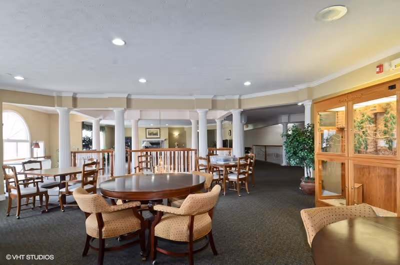 A spacious interior common area with multiple round and rectangular wooden tables surrounded by cushioned chairs. The room features white columns, large windows letting in natural light, a carpeted floor, and a wooden display cabinet with glass doors on the right side. There are plants and framed artwork in the background.