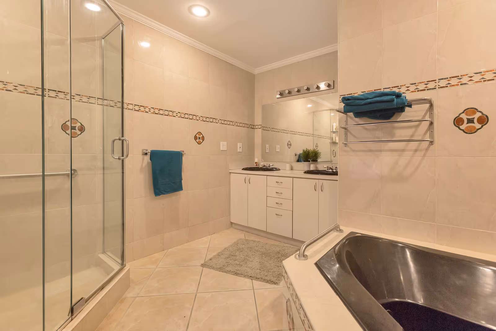 A spacious bathroom with beige tiled walls and floor. The room features a glass-enclosed shower on the left, a white vanity with two sinks and a large mirror in the center, and a dark-colored bathtub on the right. There are teal towels hanging on a towel rack and a towel bar, and a small gray rug is placed on the floor in front of the vanity. The bathroom is well-lit with recessed ceiling lights and a row of vanity lights above the mirror.