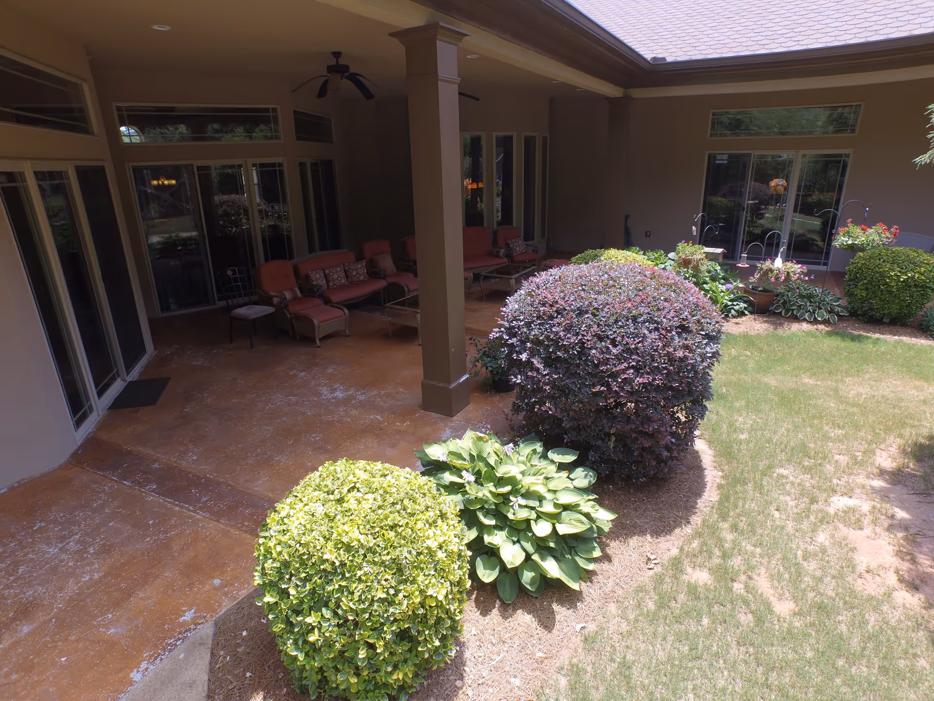 Covered outdoor patio area with cushioned seating and ceiling fans, adjacent to a garden with various shrubs and plants, and large windows and doors leading inside the building.