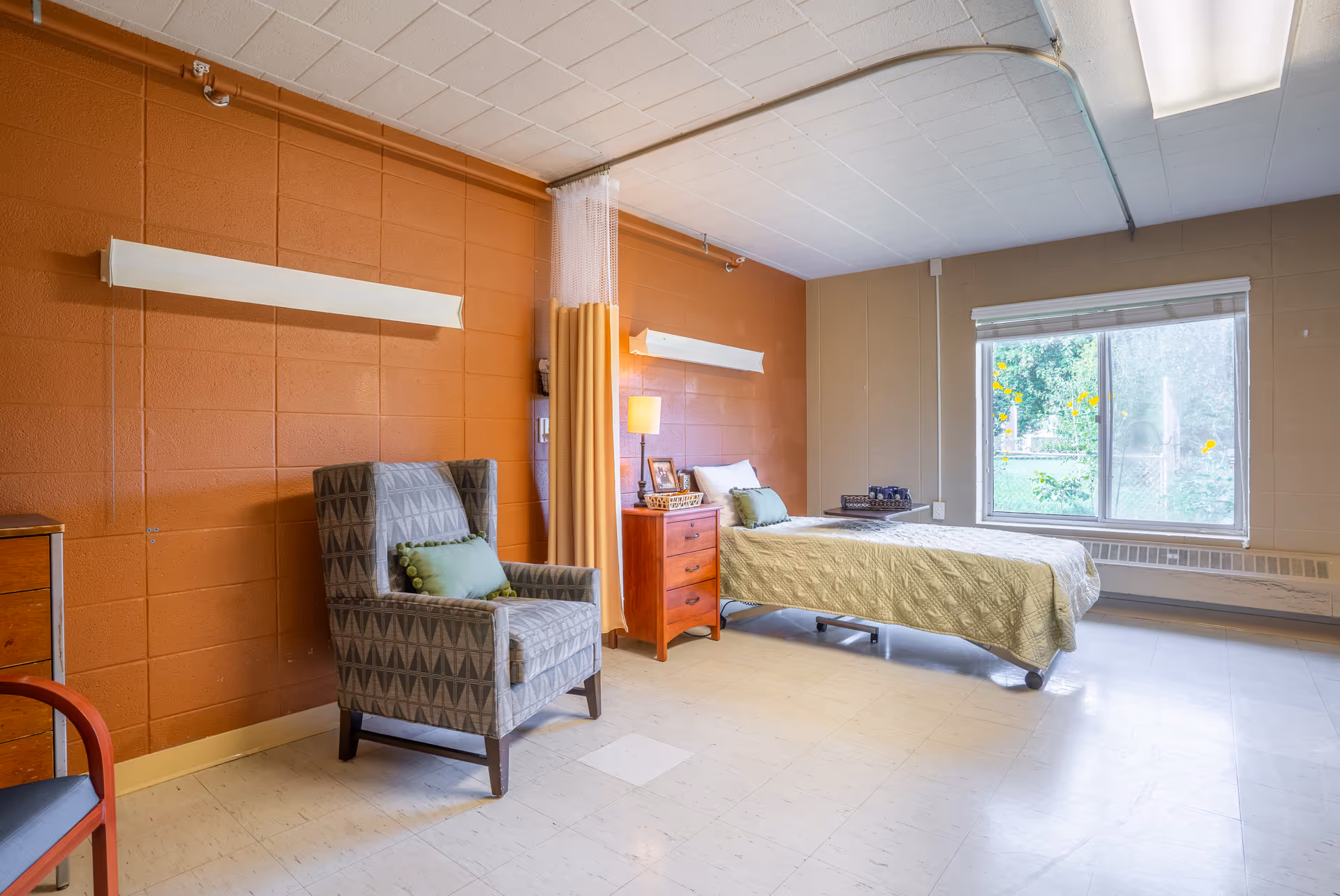 A simple, clean bedroom in a senior living facility with a single bed covered in a light green quilt, a wooden nightstand with a lamp and framed photos, a patterned armchair with a green cushion, and a large window letting in natural light. The walls are painted orange and beige, and there is a privacy curtain on a ceiling track.