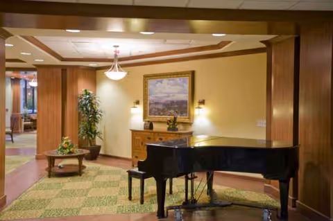 A grand piano sits in a warmly lit lobby with a painting, plants, and furniture.