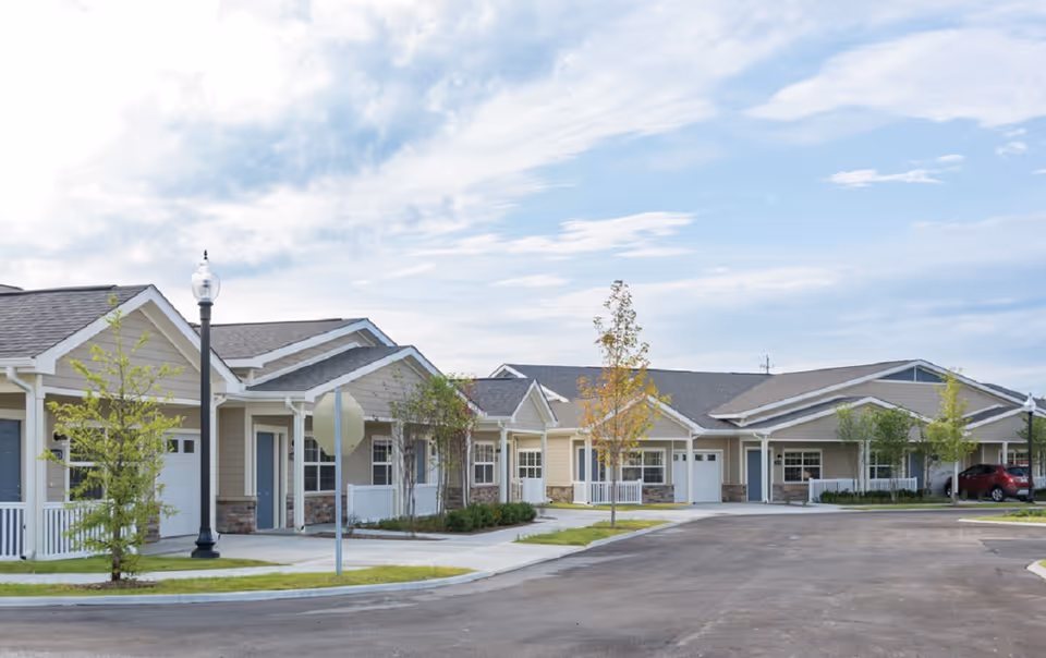Exterior view of a single-story residential building complex with beige siding, white trim, and small front porches. There are young trees planted along the sidewalk and a paved road in front. The sky is partly cloudy.