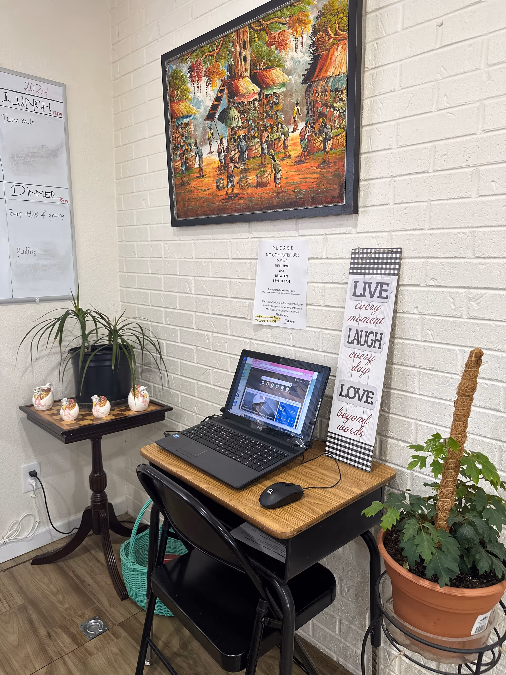 A small computer desk with a laptop and mouse on it, placed against a white brick wall. Above the desk is a colorful painting depicting a village scene with people and huts. To the right of the desk is a tall potted plant, and to the left is a small side table with four decorative ceramic chickens and a green basket underneath. A whiteboard on the left wall displays a lunch and dinner menu. A sign above the laptop requests no computer use during meal times. A decorative sign next to the desk reads 'LIVE every moment LAUGH every day LOVE beyond words.'