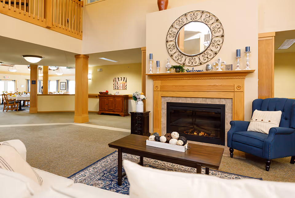 A cozy senior living common area featuring a lit fireplace with a wooden mantel decorated with candles, plants, and a round ornate mirror above it. There is a blue upholstered armchair with a white pillow to the right and a wooden coffee table with decorative balls on a tray in front. The room has beige walls, carpeted floors, and an open layout leading to a dining area with tables and chairs in the background.