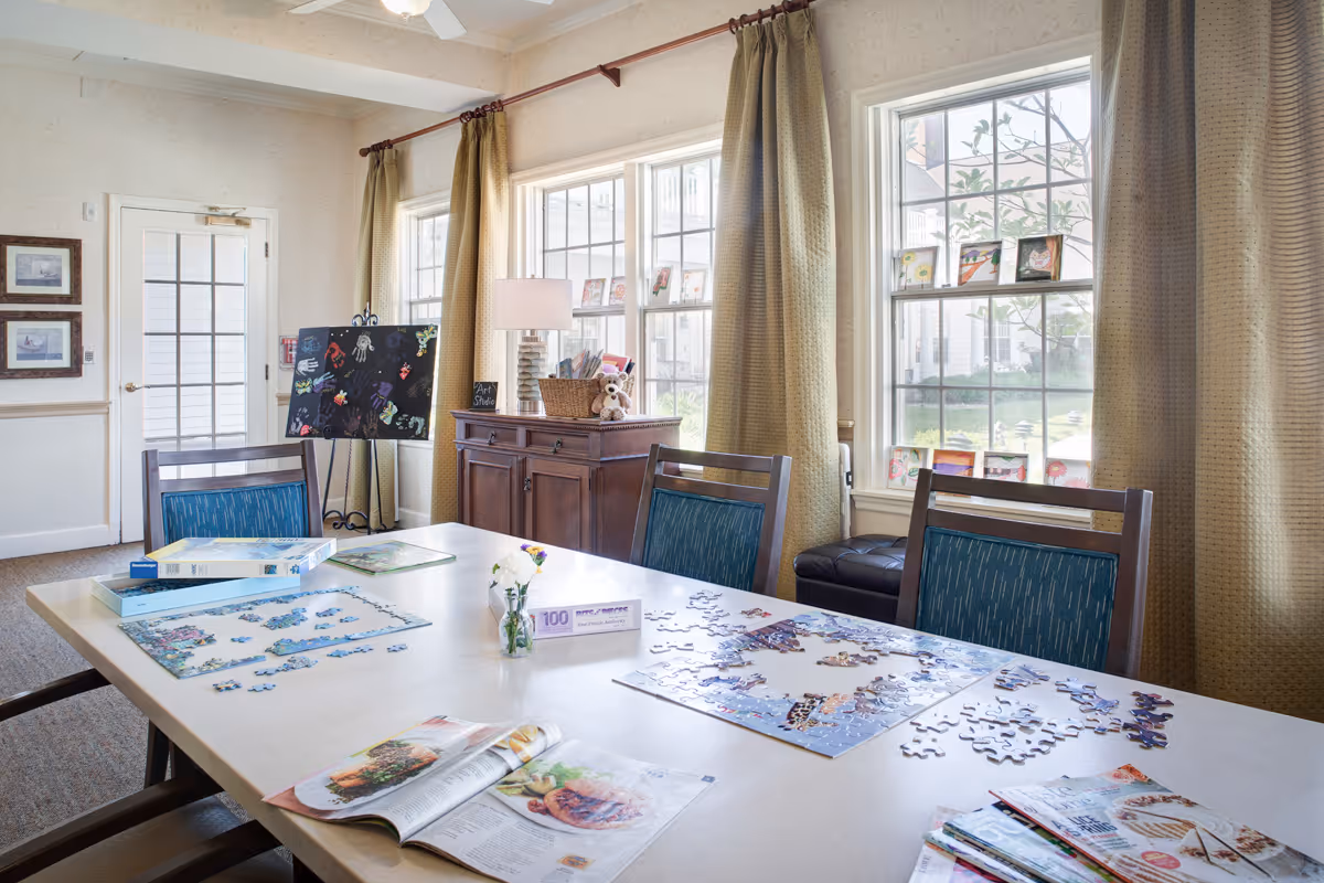 A bright room with a large table covered with partially completed jigsaw puzzles, magazines, and a small vase with flowers. The room has three windows with beige curtains, a wooden cabinet with a lamp and a basket on top, and a door with glass panels. There is an easel with artwork and framed pictures on the wall.