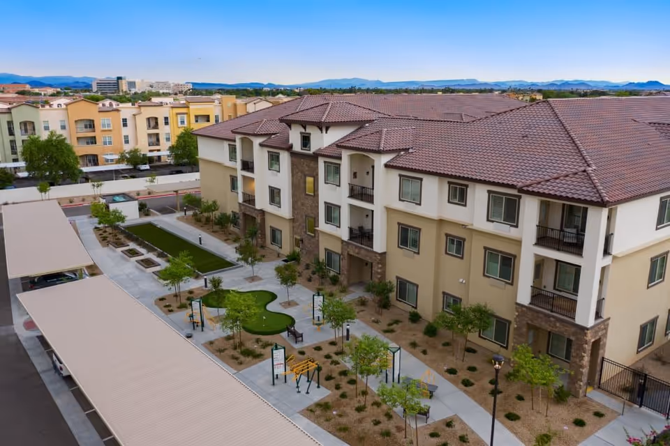 Aerial view of a multi-story residential building with beige and white exterior walls and a red-tiled roof. The building is surrounded by landscaped pathways, small trees, benches, and outdoor exercise equipment. Covered parking spaces are visible on the left side, and distant mountains can be seen in the background under a clear blue sky.