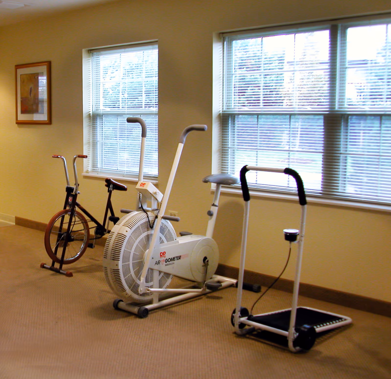 A small exercise room with three pieces of fitness equipment including a stationary bike, an air resistance exercise bike, and a small treadmill. The room has beige walls, carpeted floor, two large windows with blinds, and a framed picture on the wall.