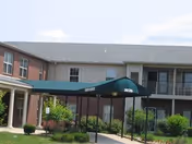 Exterior view of a two-story senior apartment building with a covered walkway entrance labeled 3020. The building has brick and siding walls, multiple windows, and a balcony on the second floor. There is greenery and shrubs around the entrance area.
