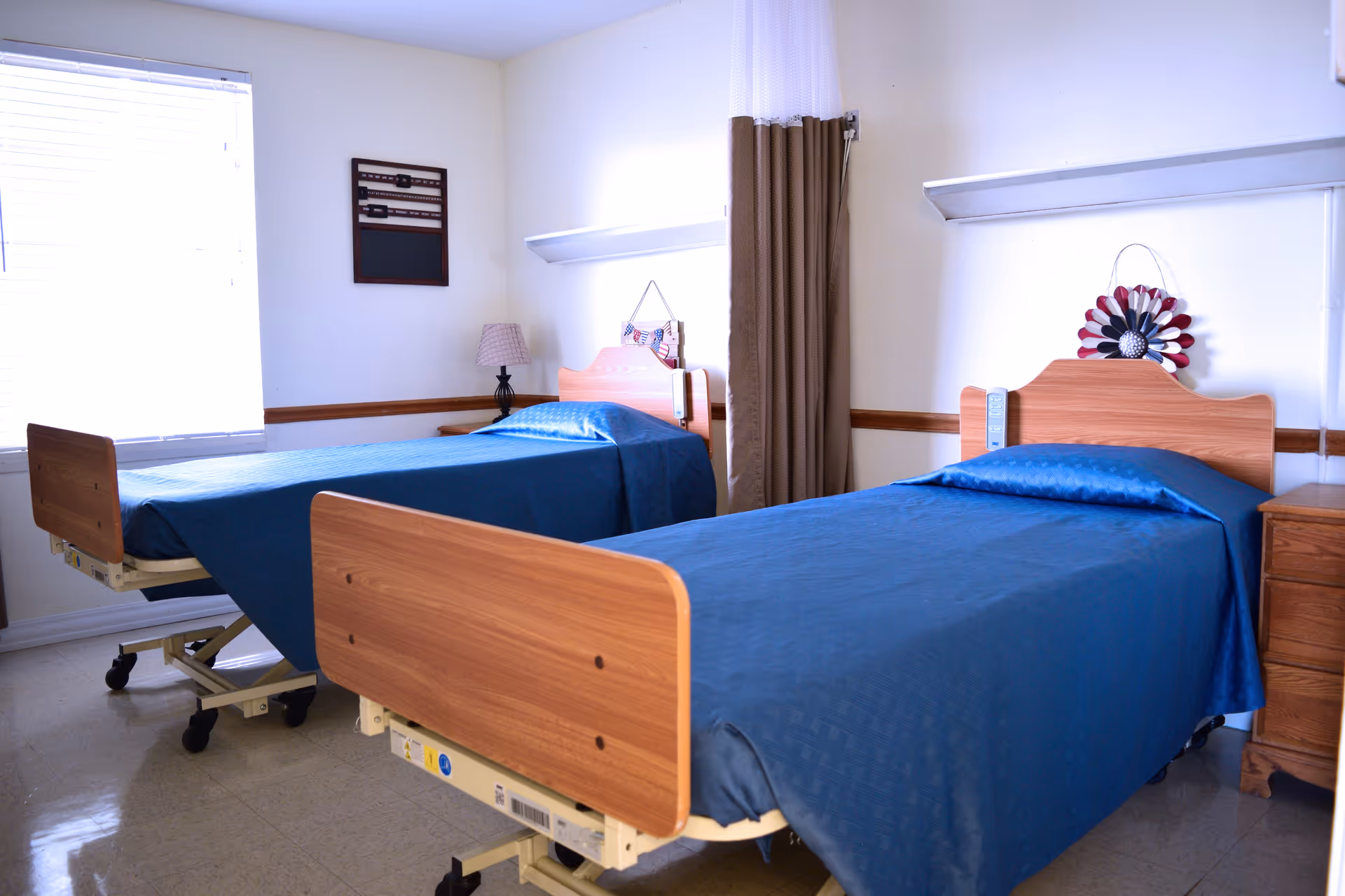 A room with two hospital-style beds with wooden headboards and footboards, each covered with blue bedspreads. There is a window with blinds on the left wall, a small nightstand with a lamp between the beds, and a privacy curtain hanging between the beds. The walls are light-colored, and there is a decorative item hanging above each bed.