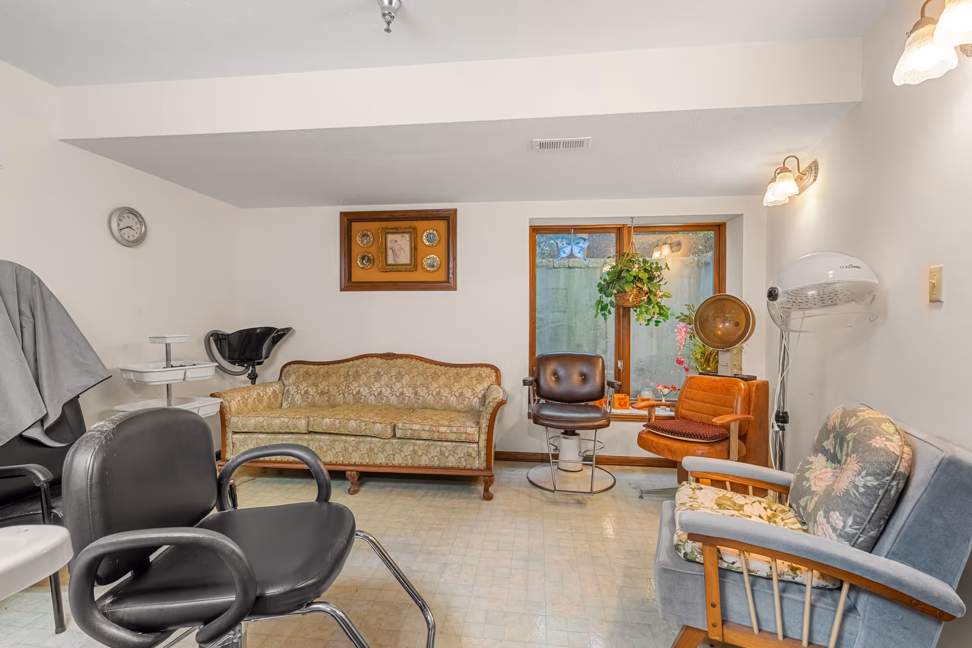 Room with a vintage sofa and various salon chairs, a hooded hair dryer, framed art, and a window with a hanging plant.