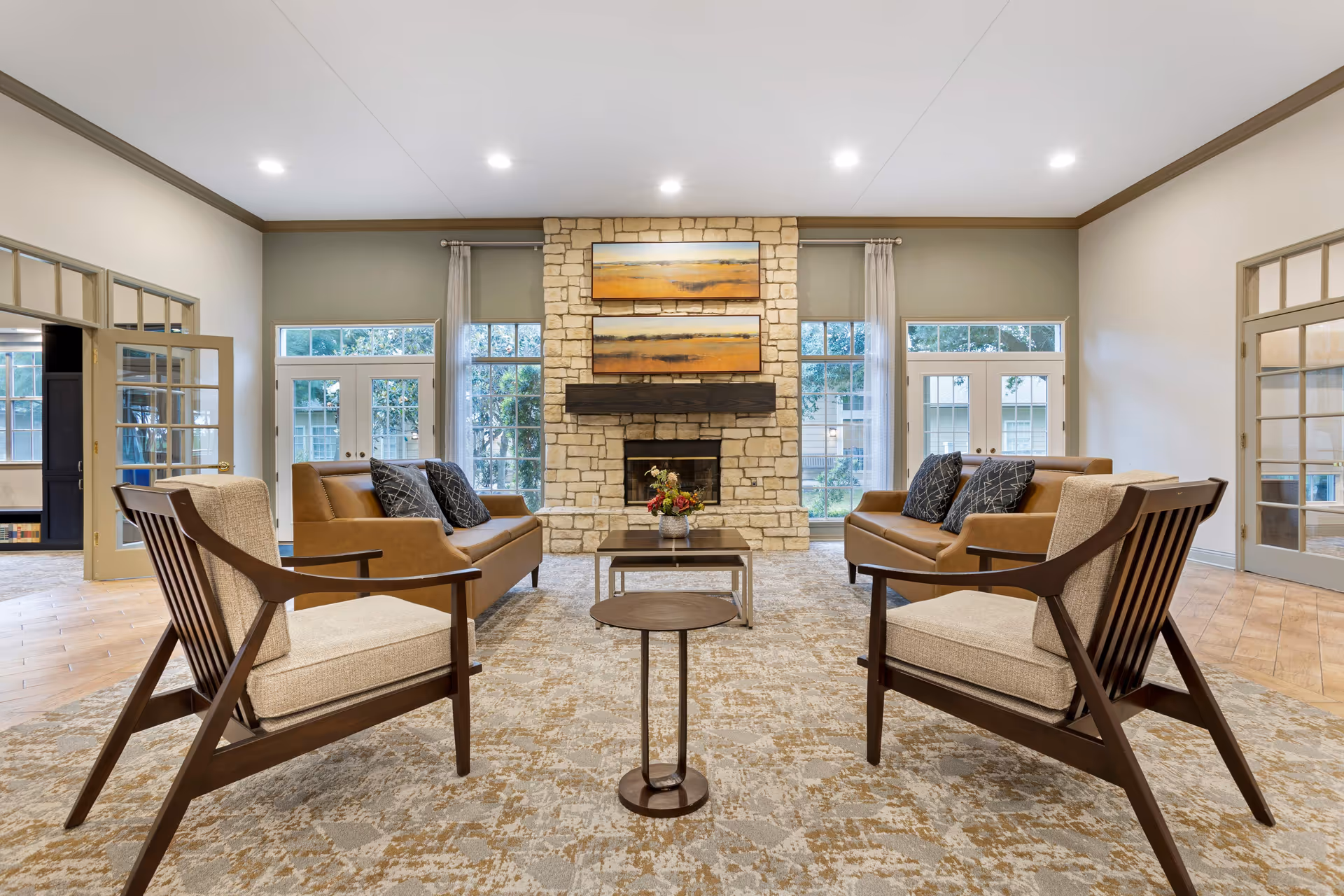 A spacious and well-lit living room in Windermere Estates Senior Living featuring two beige cushioned wooden armchairs in the foreground, two brown leather sofas with dark patterned pillows facing each other, a stone fireplace with a wooden mantle in the center, and two landscape paintings above the fireplace. Large windows and glass doors with light curtains flank the fireplace, allowing natural light to fill the room. The floor is covered with a patterned carpet, and there are two small tables in the center, one with a flower arrangement.