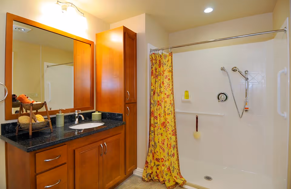 Bathroom with wooden cabinets and a black granite countertop with a sink. A large mirror is mounted above the sink. There is a shower area with a yellow floral shower curtain and a handheld showerhead. The walls are light-colored and the floor has a tiled pattern.