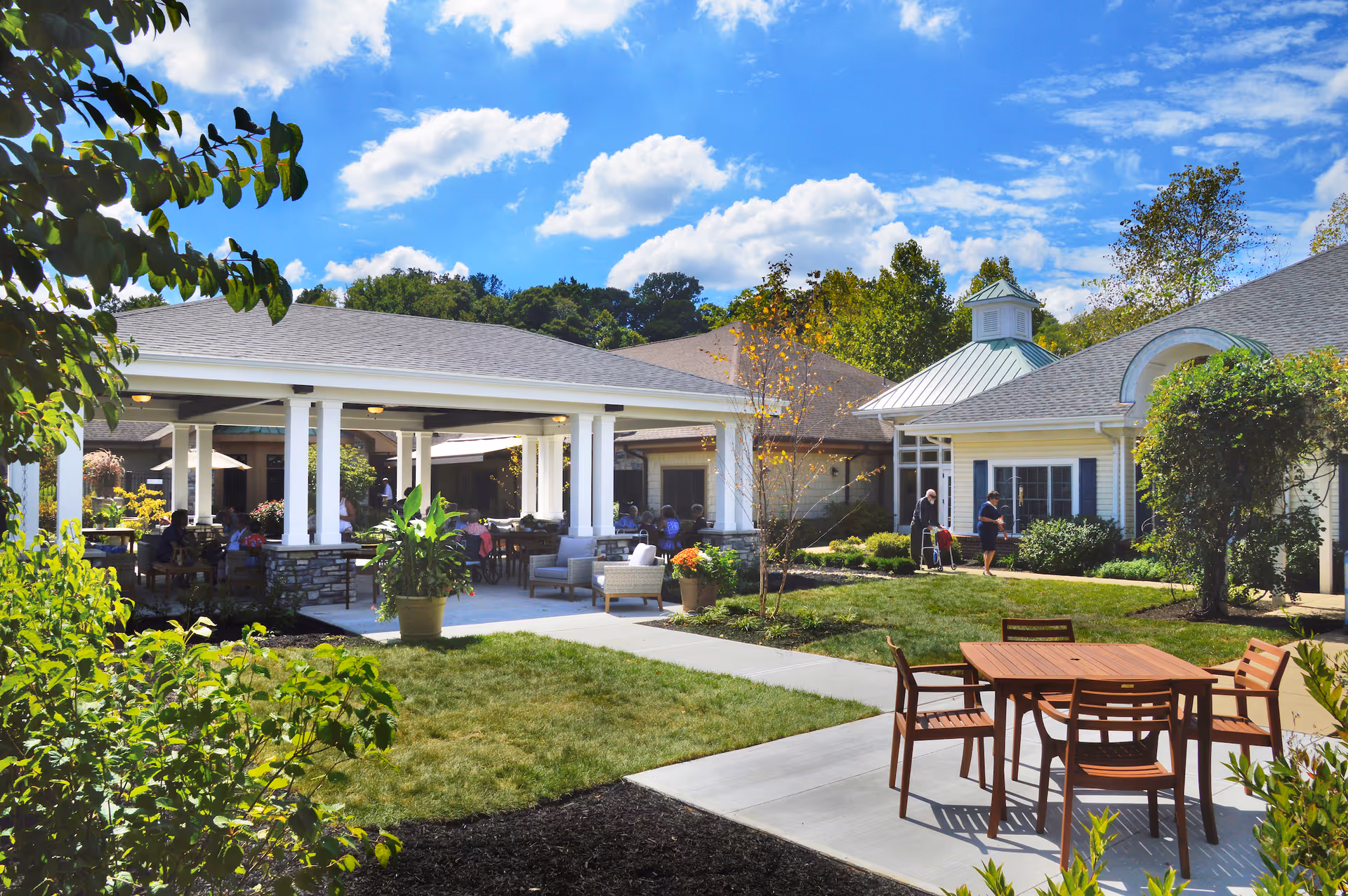 Outdoor patio area at Deupree House with a covered seating area featuring chairs and tables, surrounded by greenery and trees under a blue sky with scattered clouds. There are a few people visible in the background near the building.