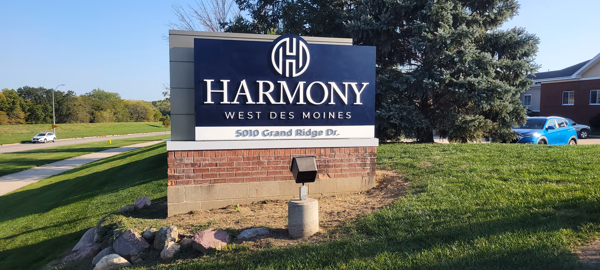 Outdoor entrance sign reading 'Harmony West Des Moines' on a brick base in a grassy area with trees and a building in the background.