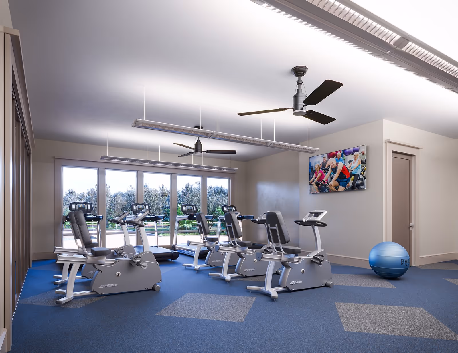 A fitness room with several exercise bikes and treadmills facing large windows that show greenery outside. The room has blue and gray patterned carpet, two ceiling fans, and a wall-mounted TV displaying a group of elderly people exercising. A blue exercise ball is placed near a closed door.