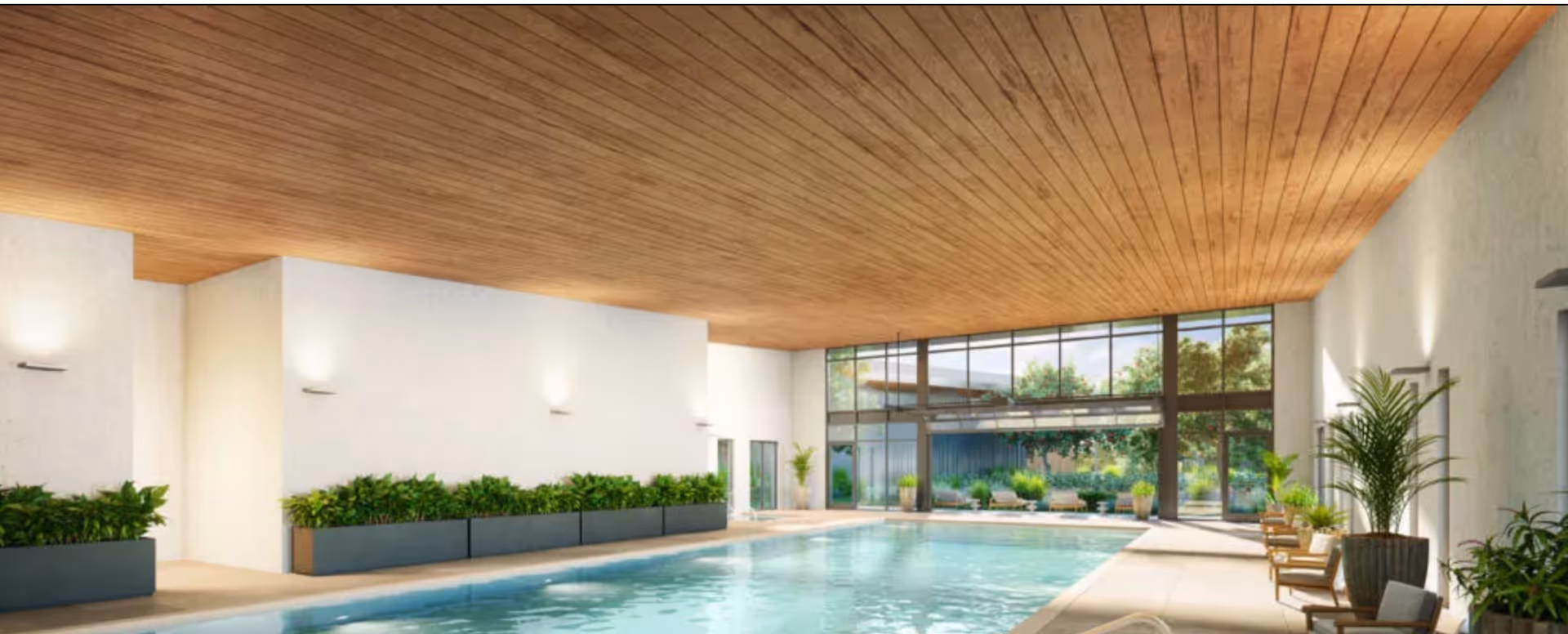 Indoor swimming pool area with a wooden ceiling, white walls, and large floor-to-ceiling windows letting in natural light. The pool is surrounded by planters with green plants and several chairs with cushions along the right side. Outside the windows, trees and outdoor seating are visible.