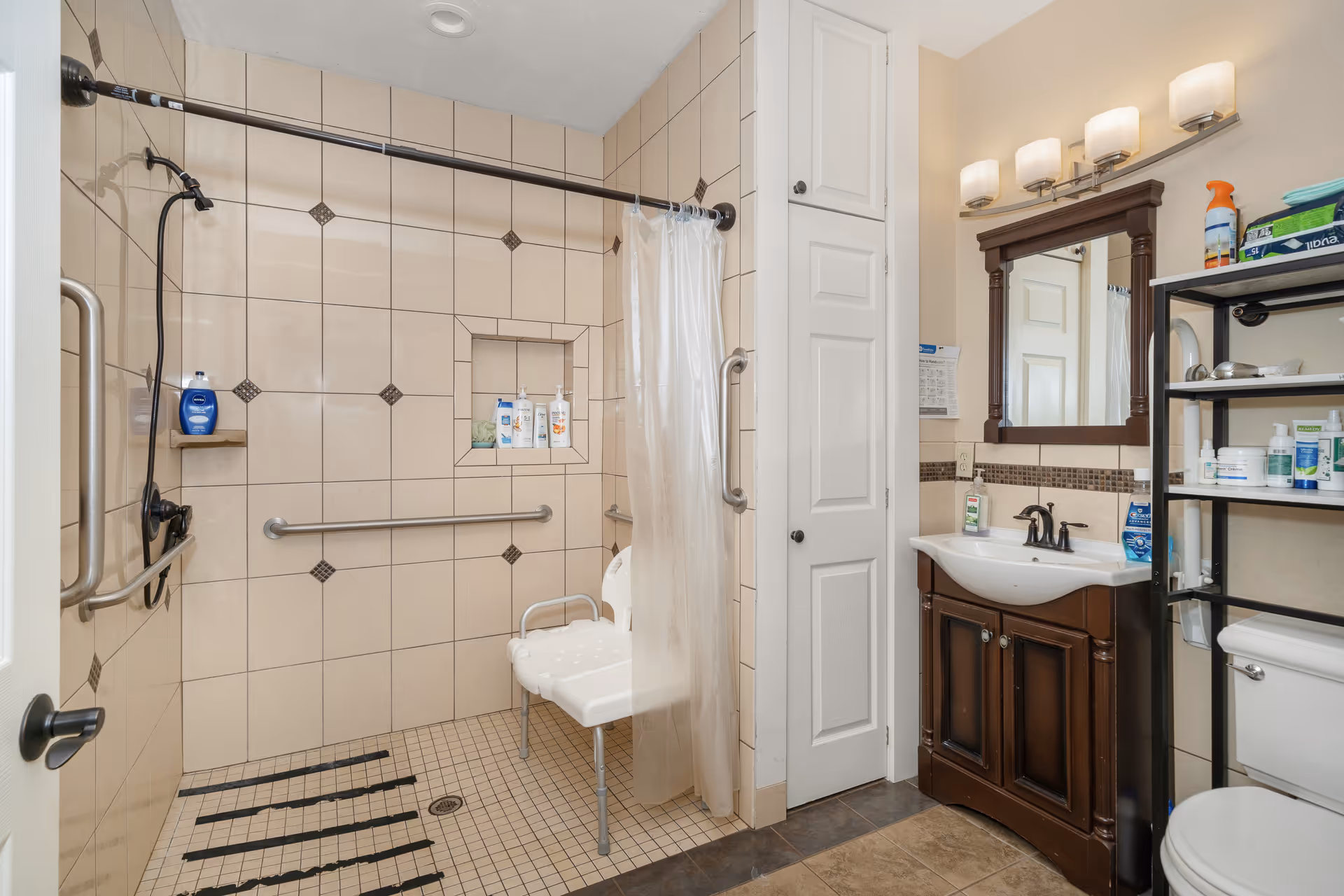 A bathroom with beige tiled walls and floor, featuring a walk-in shower with a white shower chair, grab bars, and a handheld showerhead. There is a white shower curtain on a curved rod. Next to the shower is a dark wood vanity with a white sink, a mirror with a dark wood frame, and a light fixture with three lights above it. A white tall cabinet is beside the vanity, and a black shelving unit holds various toiletries above a white toilet.