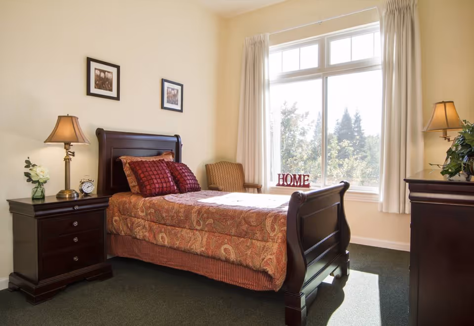 A cozy bedroom with a dark wooden bed frame and a patterned bedspread with matching pillows. There is a nightstand with a lamp, a clock, and a small vase with a white flower on the left side of the bed. A cushioned chair is placed near a large window with white curtains, and the word 'HOME' is displayed on the windowsill. A dresser with a lamp and a plant is on the right side of the room. The walls are light-colored, and two framed pictures hang above the bed.