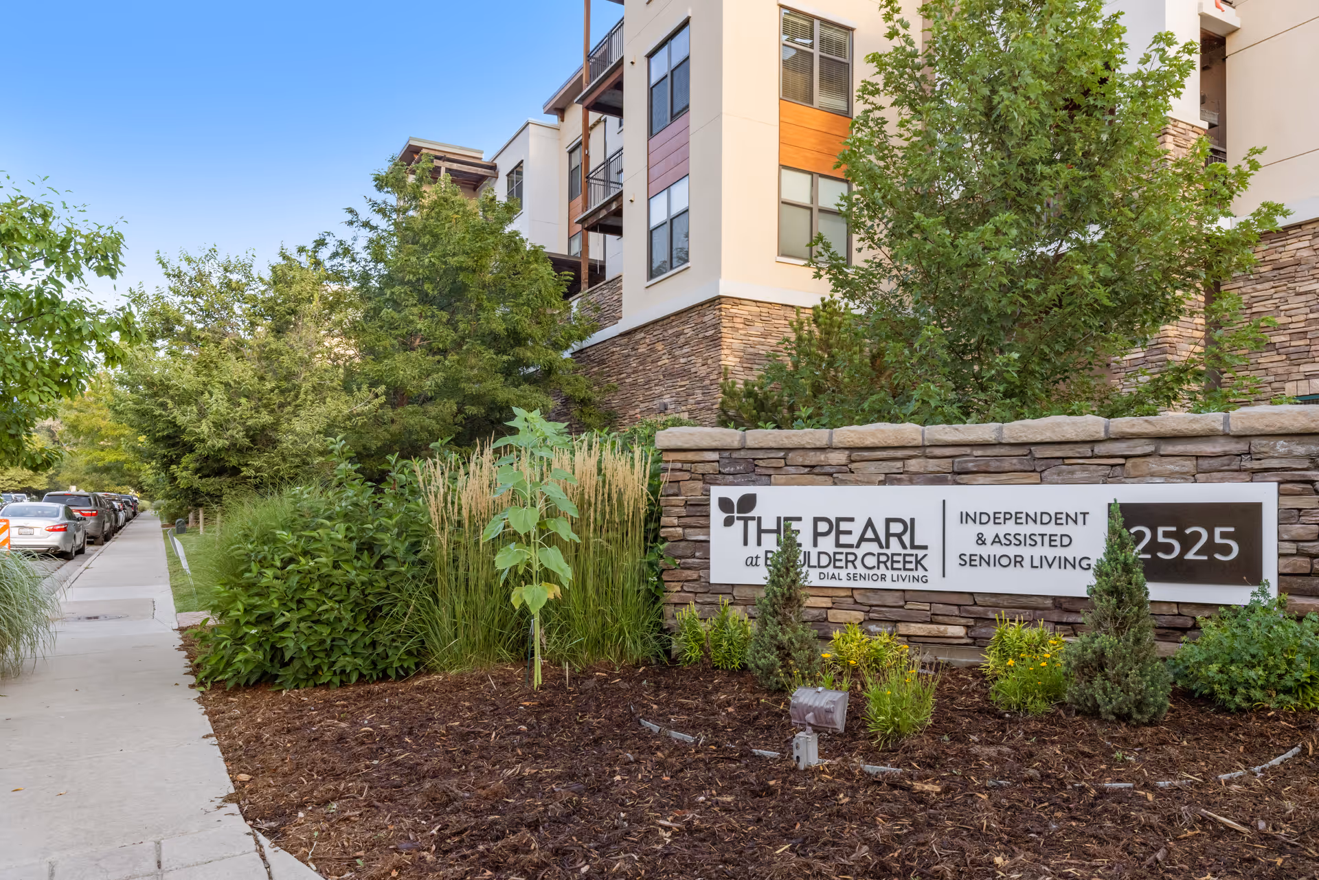 Stone entry sign and landscaped front of The Pearl at Boulder Creek senior living building along a sidewalk.