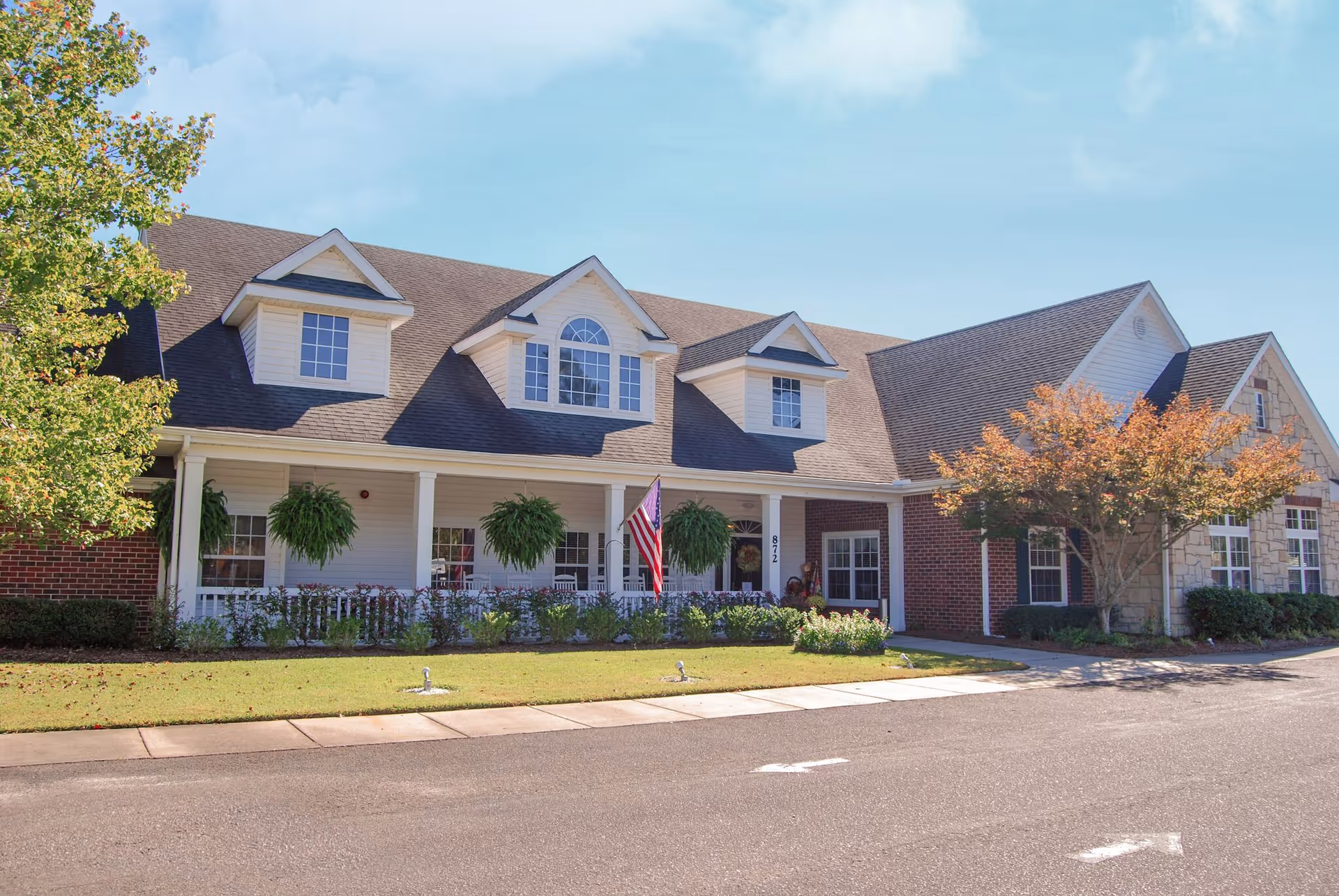 Front exterior of a single-story senior living building with a covered porch, hanging plants, American flag, and landscaped lawn.