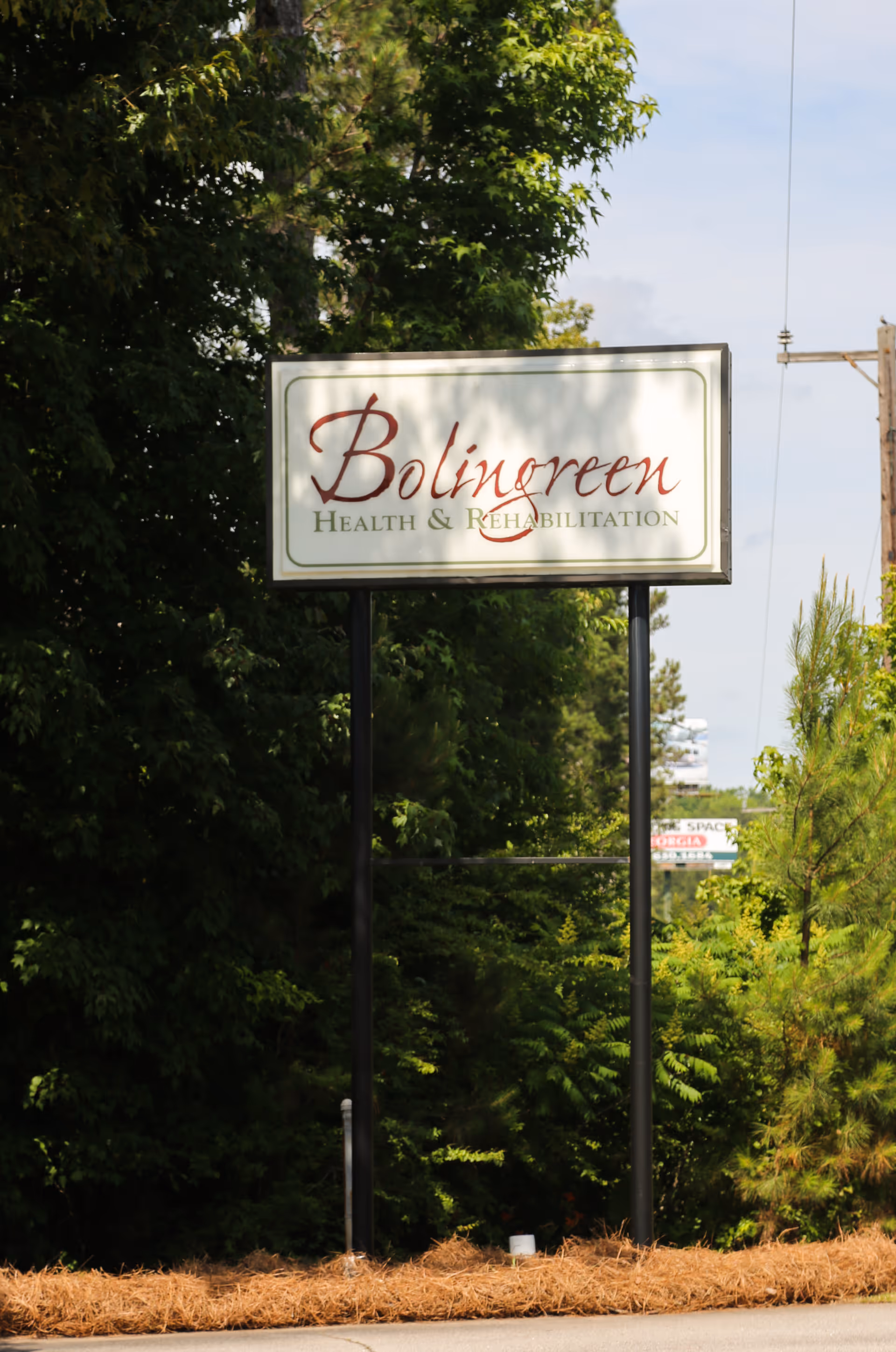 A tall sign for Bolingreen Health & Rehabilitation stands outdoors surrounded by green trees and bushes under a partly cloudy sky.