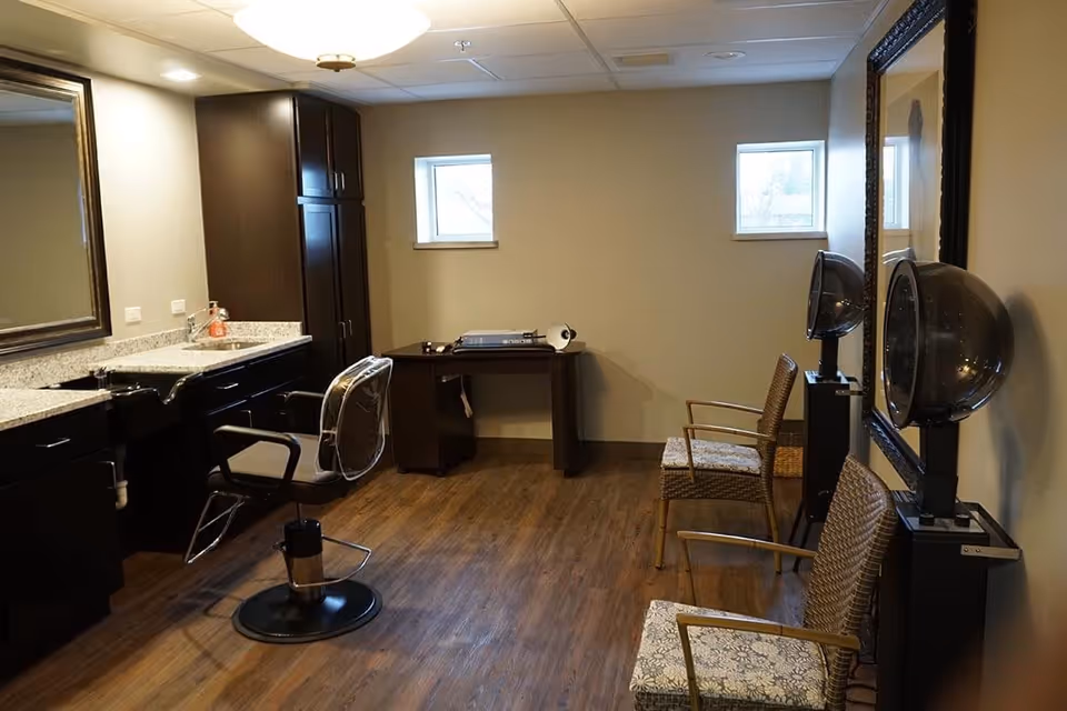 Interior view of a hair salon area in an assisted living facility featuring a styling chair in front of a large mirror and countertop with a sink. On the opposite side, there are two hair drying stations with chairs and hooded hair dryers. The room has wooden flooring, two small windows, and a desk with a lamp in the corner.