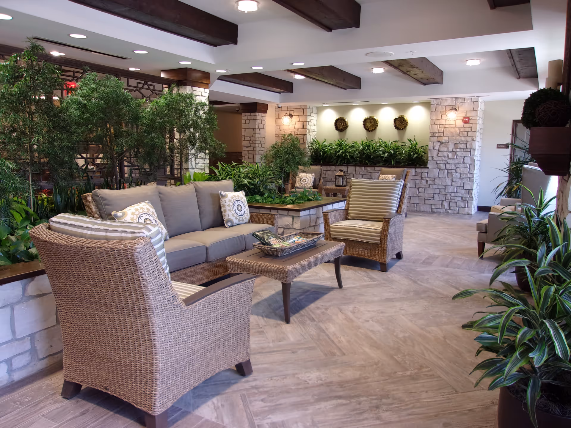 A cozy indoor seating area with wicker furniture including a sofa and two armchairs with cushions, a coffee table with magazines, surrounded by various green plants and stone wall accents under a ceiling with exposed wooden beams and recessed lighting.
