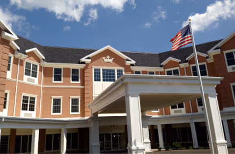 Exterior view of a multi-story brick building with white trim and a covered entrance. An American flag is flying on a flagpole in front of the building under a partly cloudy blue sky.