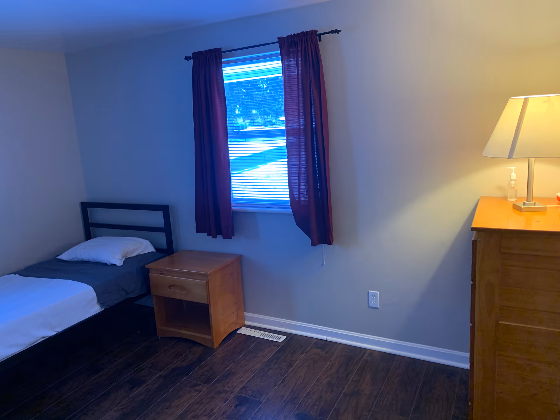 Small bedroom with a single bed, wooden nightstand and dresser, a window with burgundy curtains, and a lamp.