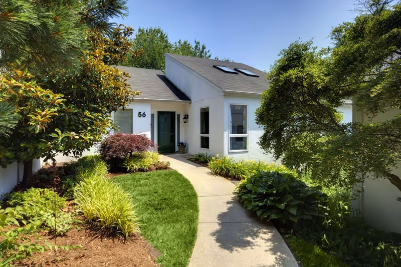 A white single-story building with a sloped roof and skylights, surrounded by lush green trees and shrubs. A curved concrete pathway leads to a green front door marked with the number 56. The garden area is well-maintained with various plants and bushes under a clear blue sky.