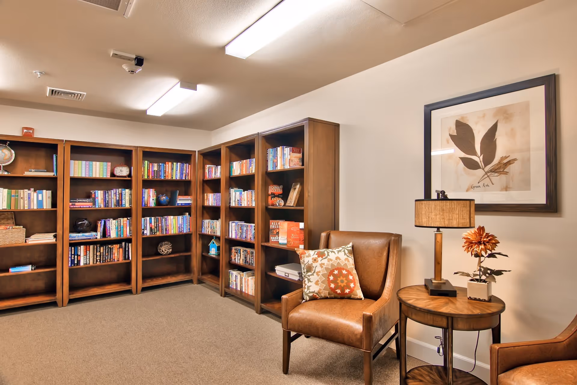 A cozy reading room with wooden bookshelves filled with books along the walls. There are two brown leather armchairs with patterned cushions, a round wooden side table with a lamp and a small flower arrangement. A framed botanical artwork hangs on the wall above the table.