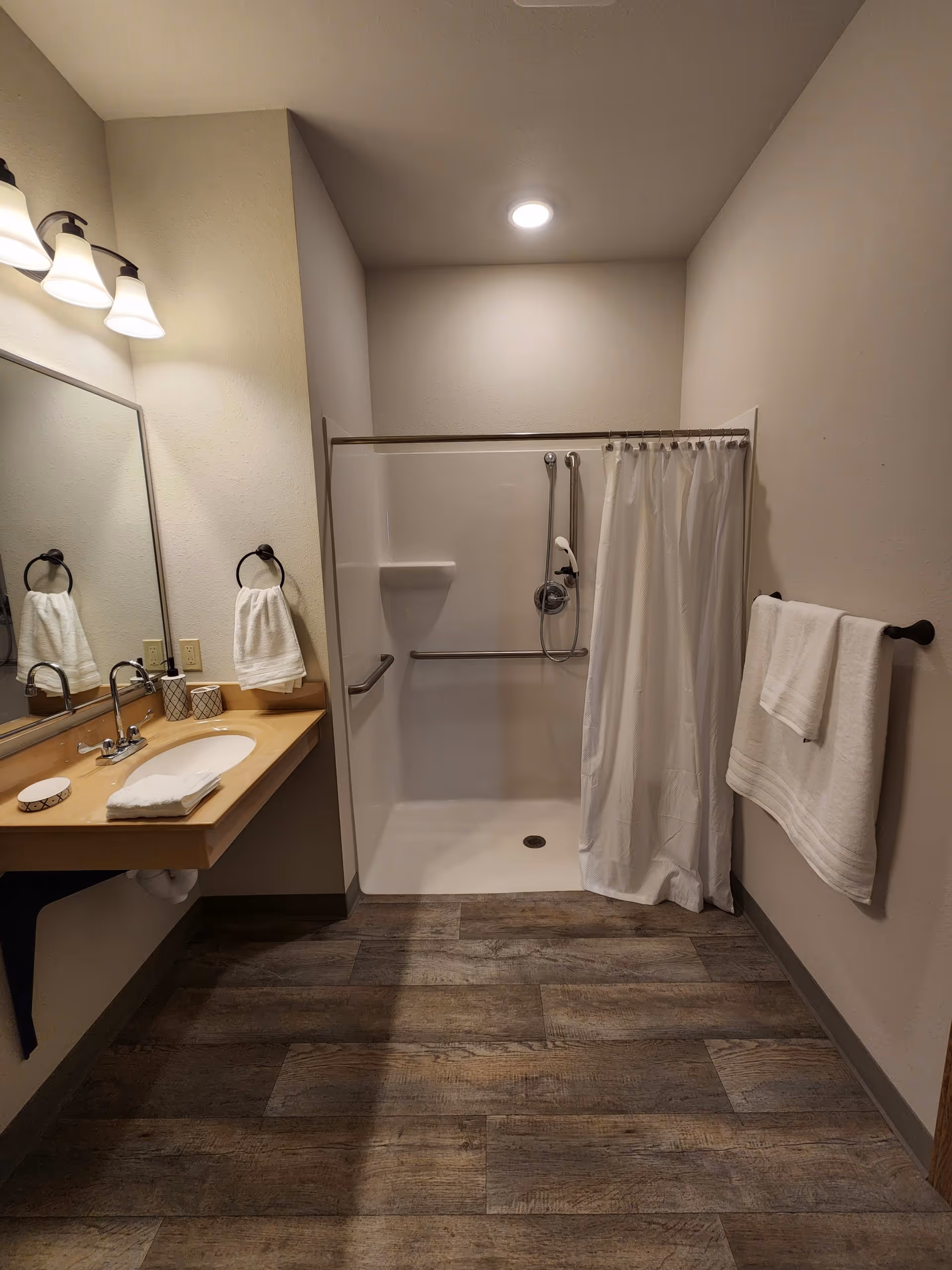 A senior living facility bathroom with a walk-in shower featuring grab bars and a handheld showerhead. There is a white shower curtain partially drawn to the side. To the left, a wooden countertop with a sink, faucet, soap dispenser, and folded white towels is visible. Above the sink is a large mirror and three light fixtures. A towel ring with a white towel is mounted on the wall beside the sink, and another towel hangs on a towel bar on the right wall. The floor has wood-like tiles.