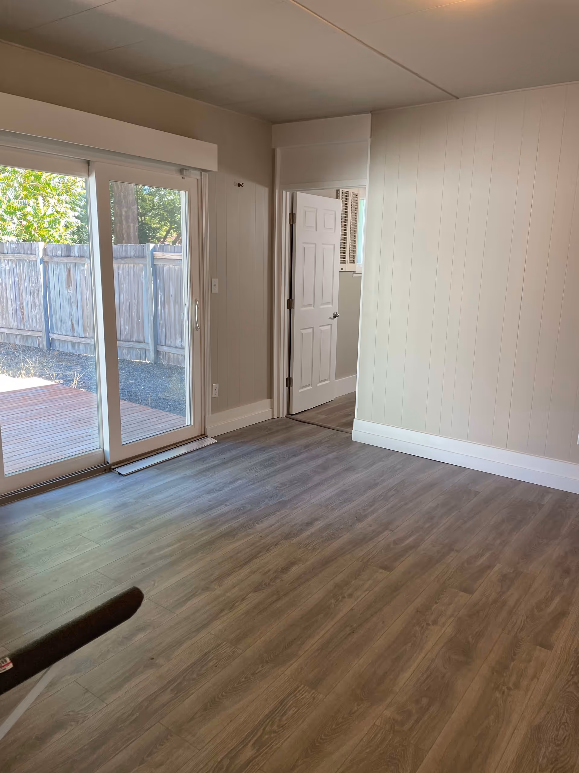 Empty room with light wood flooring, beige walls, a sliding glass door leading outside to a wooden deck and fenced yard, and an open white door leading to another room.
