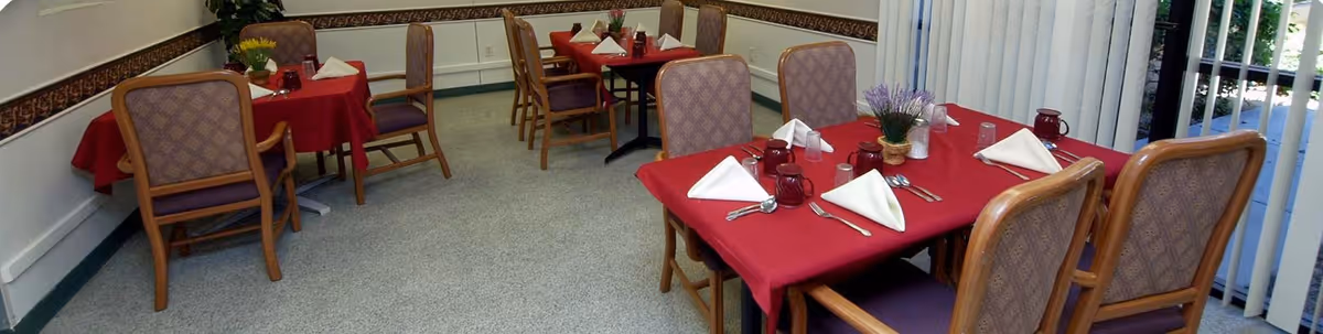 Dining room with several tables covered with red tablecloths, each set with white folded napkins, utensils, and glasses. The room has patterned chairs around the tables, a carpeted floor, and vertical blinds on the windows.