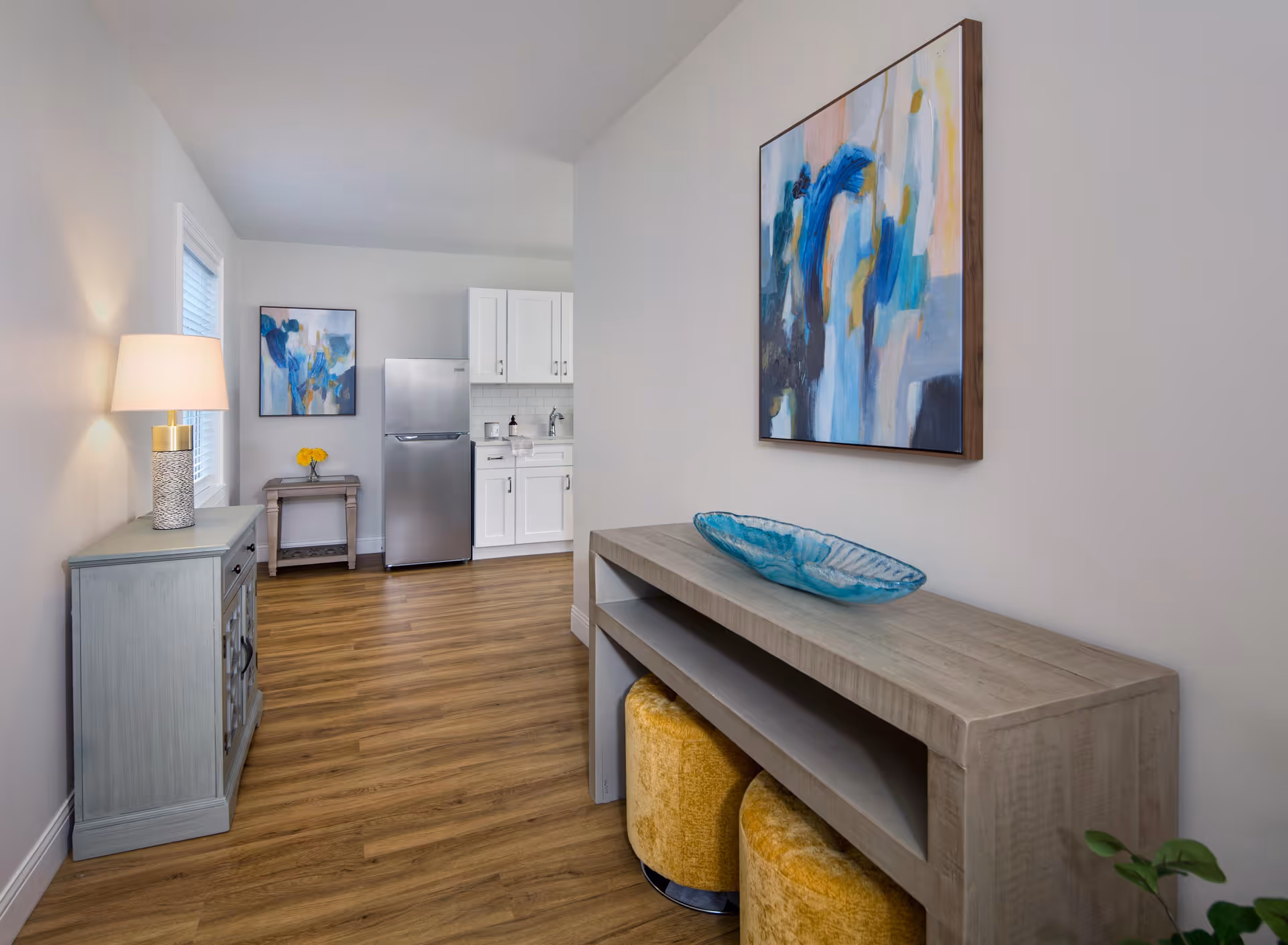 Interior view of a modern living space with light wood flooring and white walls. On the right, there is a wooden console table with a blue decorative glass bowl on top and two yellow ottomans underneath. Above the console table hangs an abstract painting with blue, white, and yellow tones. On the left side, there is a light blue cabinet with a lamp on top, next to a window with blinds. In the background, a small kitchen area with white cabinets and a stainless steel refrigerator is visible, along with a small table holding a vase with yellow flowers and another abstract painting on the wall.