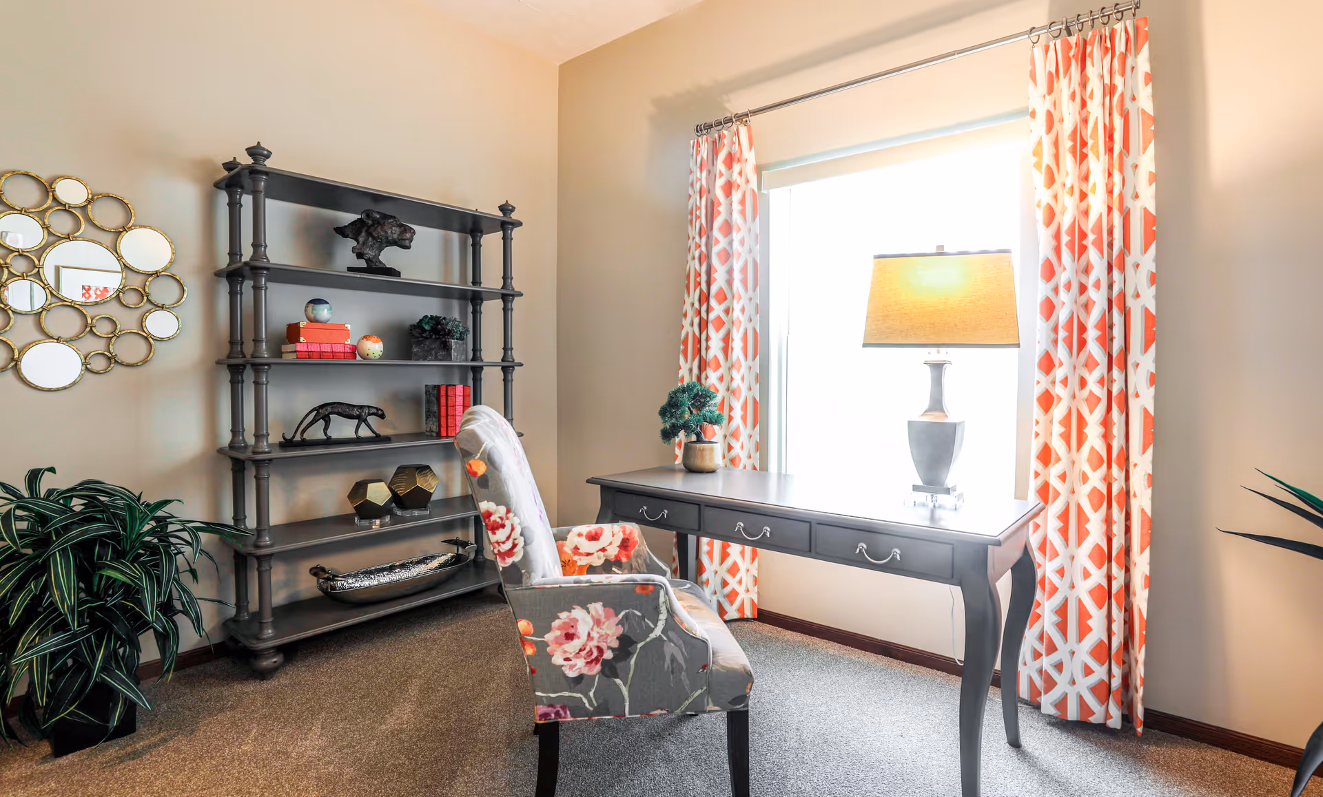 A bright room with a large window covered by orange and white patterned curtains. In front of the window is a dark wooden desk with three drawers, a table lamp, and a small potted plant. A floral upholstered chair is positioned at the desk. To the left, there is a tall dark wooden shelving unit with decorative items including books, sculptures, and vases. A round mirror with multiple small circular mirrors is mounted on the beige wall. A green potted plant is on the floor near the shelving unit.
