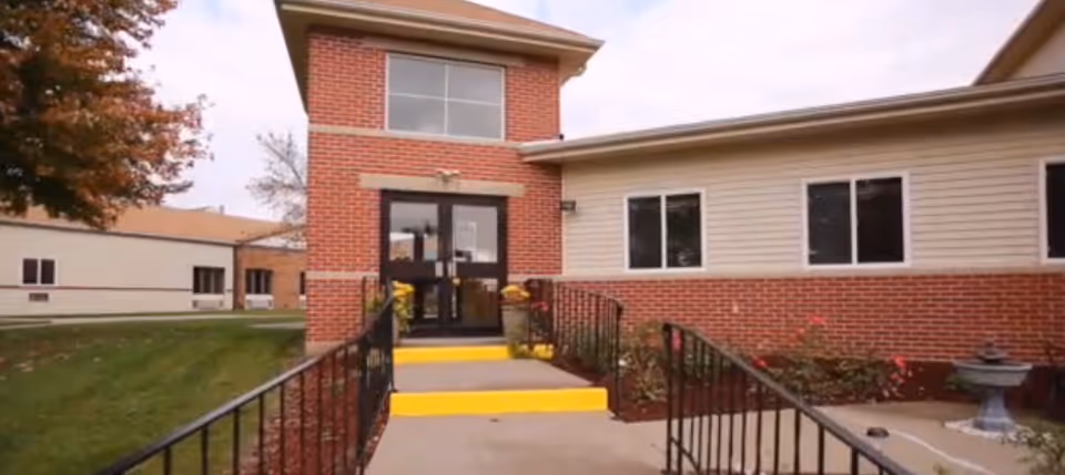 Entrance to a senior living community building with a brick and siding exterior, a concrete walkway with yellow safety markings on the steps, black metal railings, and some landscaping including a small fountain on the right side.