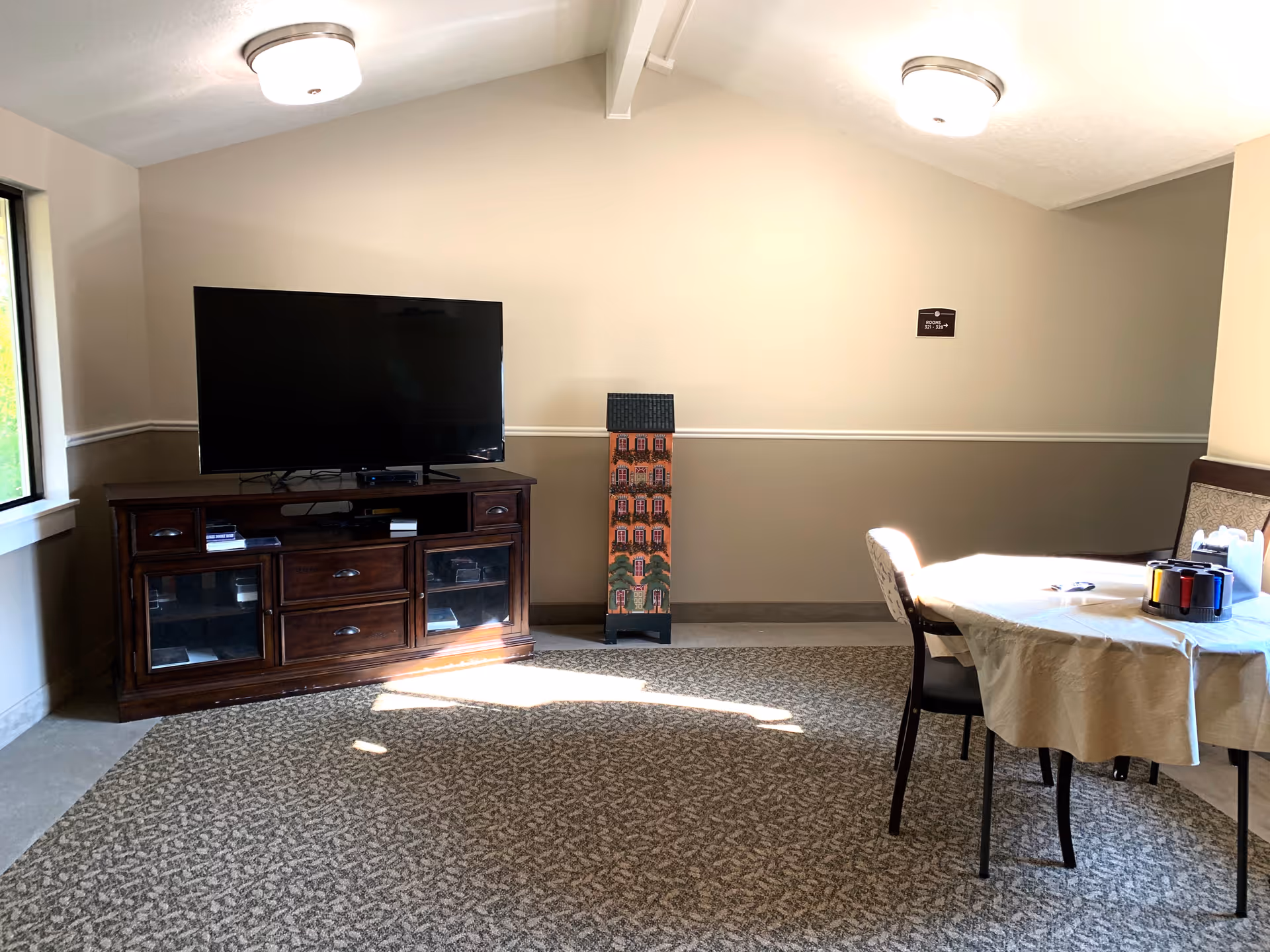 A small common area with a flat-screen TV on a wooden cabinet, a decorative tall narrow cabinet with a house design, and a round table covered with a beige tablecloth. The table has chairs around it and some items on top. The room has beige walls with a chair rail and carpeted floor.