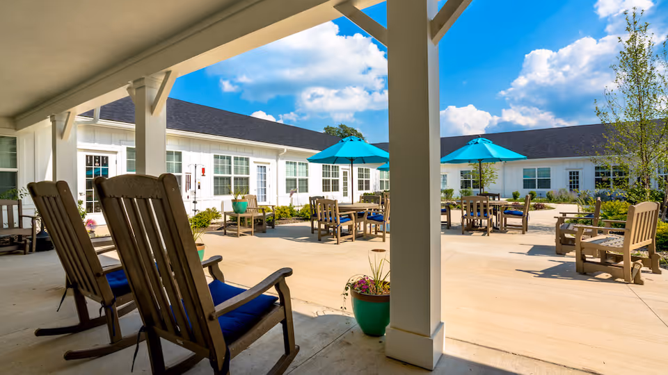 Outdoor patio area at Demaree Crossing with wooden rocking chairs and tables with blue umbrellas under a clear blue sky. The patio is surrounded by a white building with multiple windows and doors, and there are potted plants placed around the seating area.