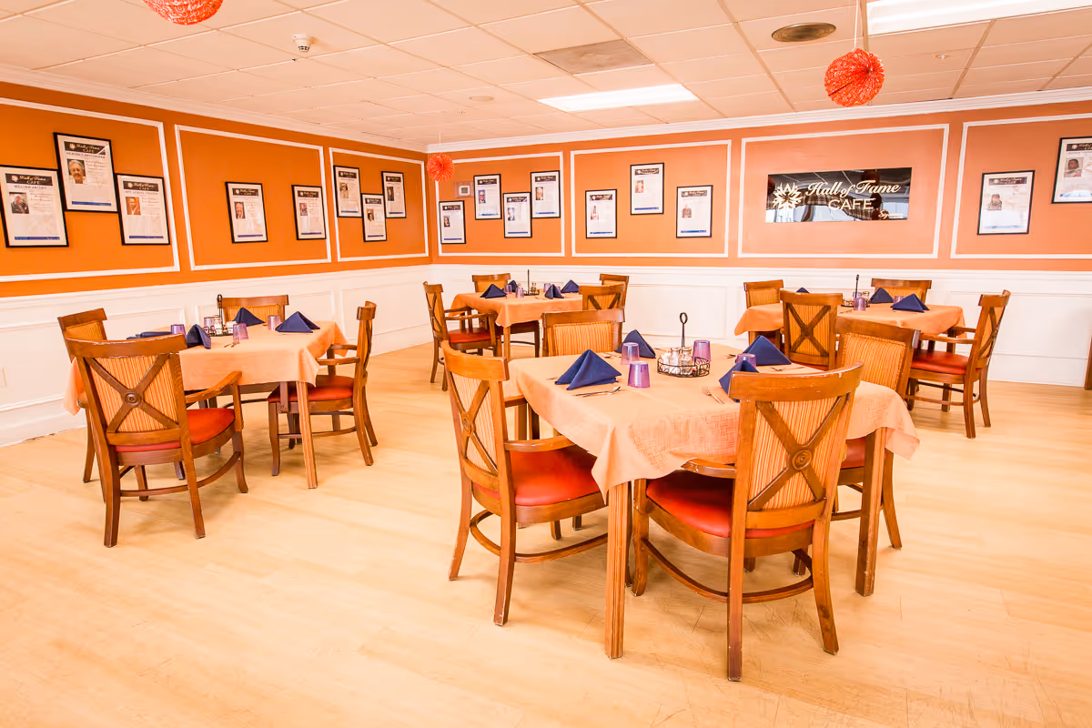 Dining room with several wooden tables covered with peach-colored tablecloths and set with blue folded napkins and purple glasses. The walls are painted orange with white trim and decorated with framed photos and articles. A sign on the wall reads 'Hall of Fame Cafe'.