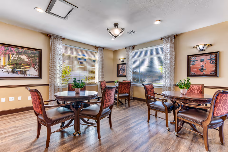 Sunny dining room with round wooden tables, upholstered chairs, potted plants, and large windows with blinds.