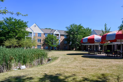 A three-story brick senior living building with a grassy courtyard, pond, and red-and-white striped outdoor canopies.