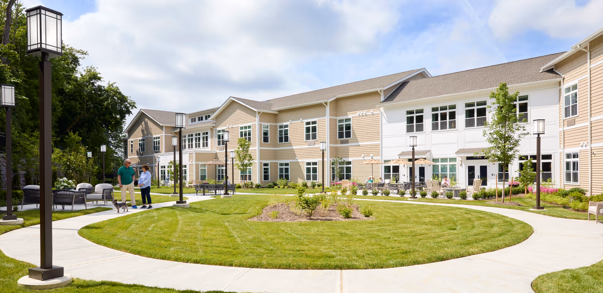 Two people walking a small dog along a circular sidewalk in the landscaped courtyard of a two-story senior living building.